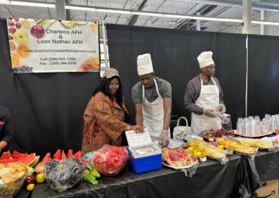 Three people are preparing food at a colorful fruit and dessert display table. Banners overhead read "Charleon AFH & Leon Nathan AFH." Various fruit platters and drinks are visible on the table.
