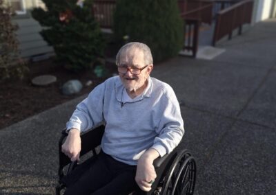 An elderly man in a wheelchair is outside on a driveway near a residential house, wearing a gray sweatshirt and dark pants.