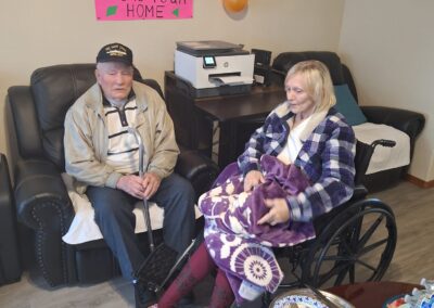 An elderly man and woman sit in a living room, the man on a sofa and the woman in a wheelchair. A welcome sign with balloons is on the wall behind them. Various items are on the coffee table.