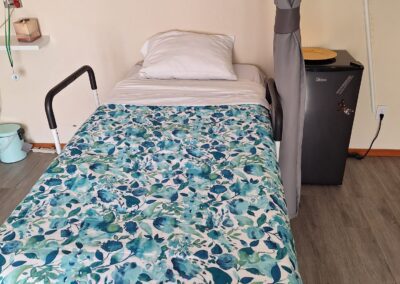 A small room with a single bed covered in blue and green floral bedding, a curtain partially drawn, a mini-fridge, and a wall-mounted picture frame above the bed.