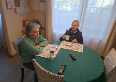 Two older adults are sitting at a dining table with cups of coffee in front of them. The table is set with placemats, a remote control, and a cordless phone. The room features a large window and a bulletin board.