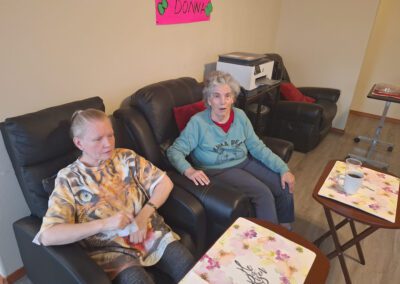Two elderly women sitting on black recliners in a room with a "Welcome Donna" sign on the wall. They have drinks on small tables in front of them.
