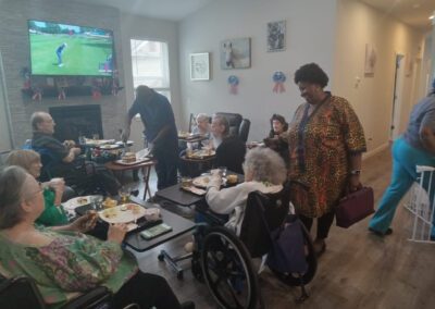 A group of people, some in wheelchairs, gather around tables having a meal in a cozy room. A TV displays a golf game. A couple of caregivers are assisting. Patriotic decorations adorn the walls.