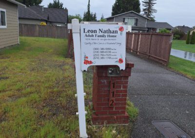 Sign for Leon Nathan Adult Family Home placed on a brick pillar outside a house, displaying contact information and surrounded by grass and houses in the background.