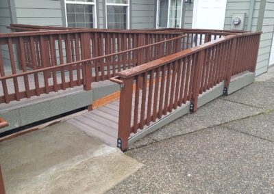 A brown wooden wheelchair ramp leading to the entrance of a gray house with white-framed windows and a white door.