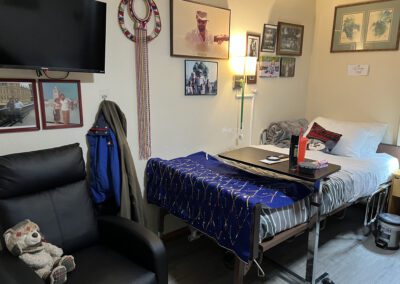 An organized room with a single bed, side table, chair, and wall-mounted TV. Personal items and photographs decorate the walls, and a teddy bear and blue jacket are placed on the chair.
