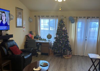A cozy living room with a Christmas tree, a ceiling fan, a TV, desk, and a leather chair. The room has hardwood flooring, large windows, and a coffee table with a bowl of snacks.